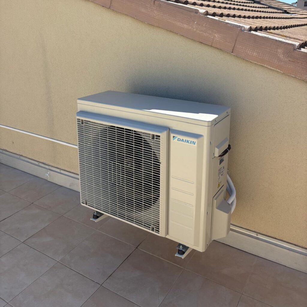 Unité extérieure de climatisation Daikin beige, posée sur un sol carrelé près d'un mur crépi sous un toit en tuiles. Le logo DAIKIN est visible.