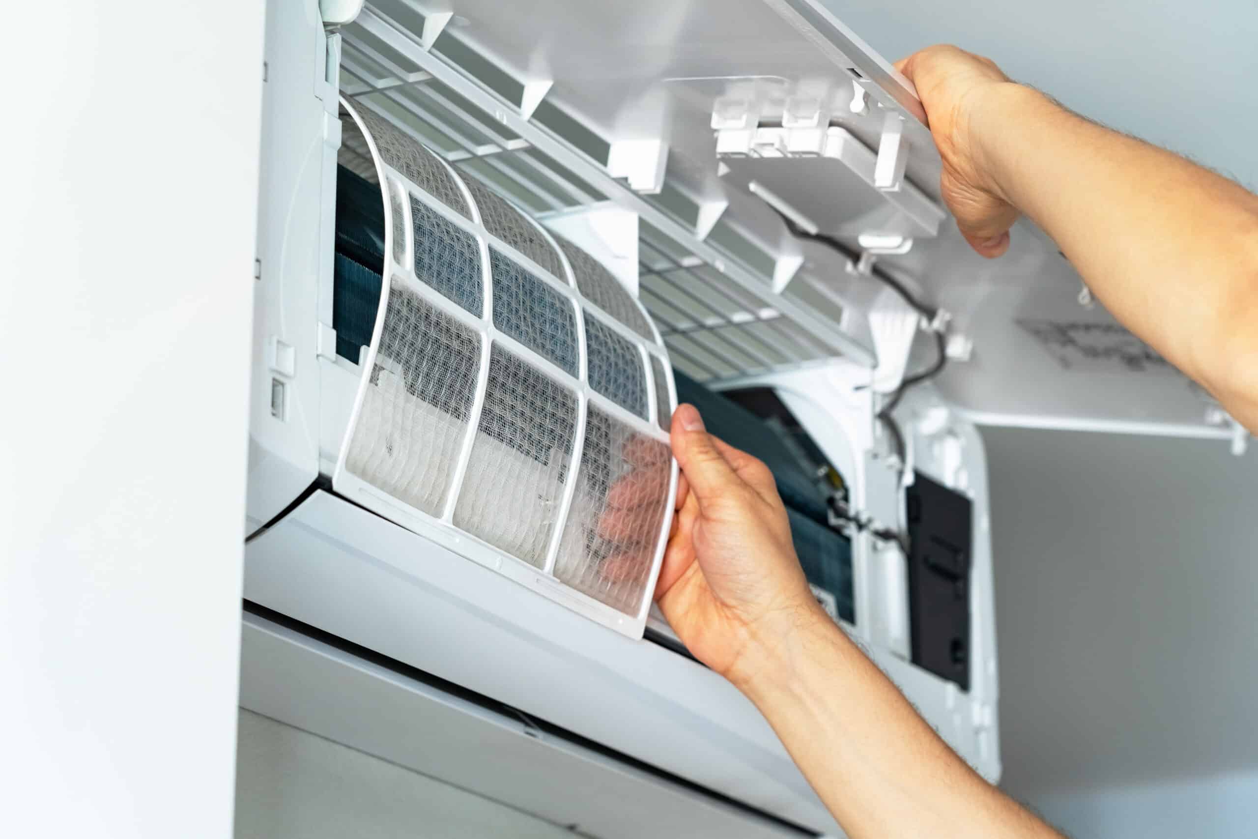 Mains retirant un filtre à air sale et poussiéreux d'un climatiseur mural blanc ouvert, montrant la nécessité d'un nettoyage.