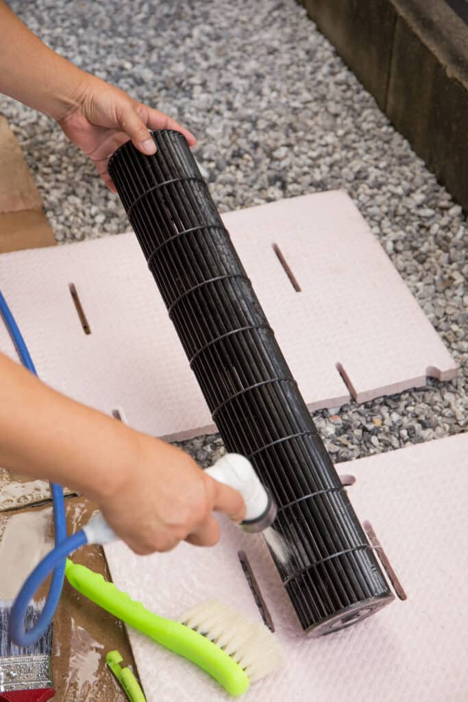 Mains nettoyant un ventilateur de climatiseur sale avec de l'eau et un pulvérisateur. La soufflante noire est rincée sur des tapis roses extérieurs.
