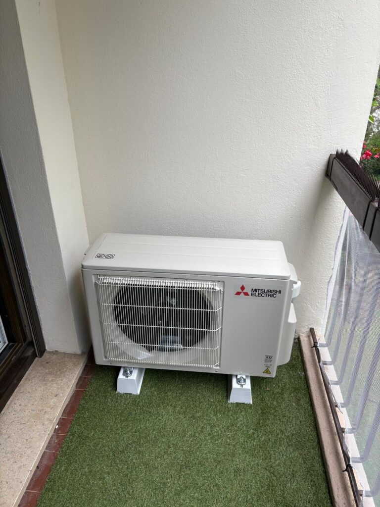 Unité extérieure Mitsubishi Electric R32 sur supports blancs, posée sur gazon artificiel d'un balcon. Mur clair, porte et garde-corps.